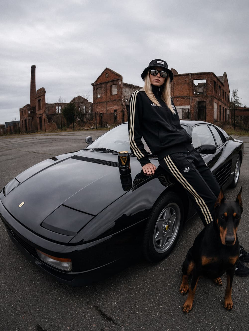 Campagna moda generata con AI: modella in tuta sportiva sul cofano di una coupé nera davanti a una fabbrica dismessa, con un dobermann accanto.